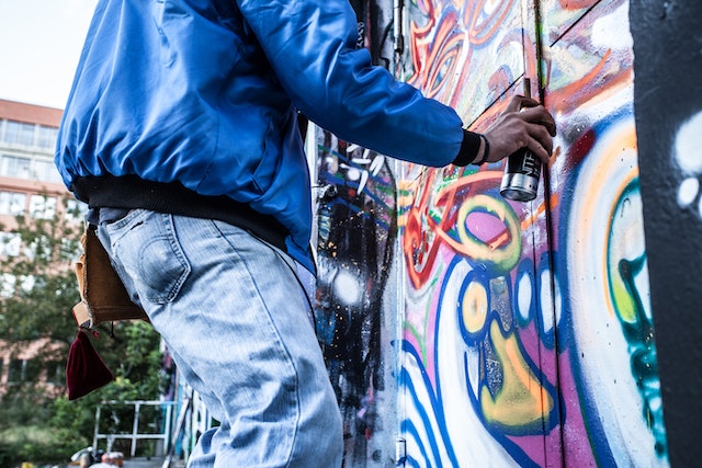 person in a blue jacket using spray paint to tag a building covered in graffiti